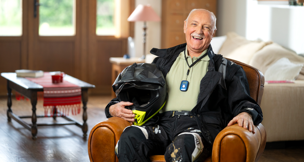 Oudere man zit lachend in een gezellige woonkamer, houdt een motorhelm vast en draagt een zwart motorpak met een medisch alarmapparaat om zijn nek.
