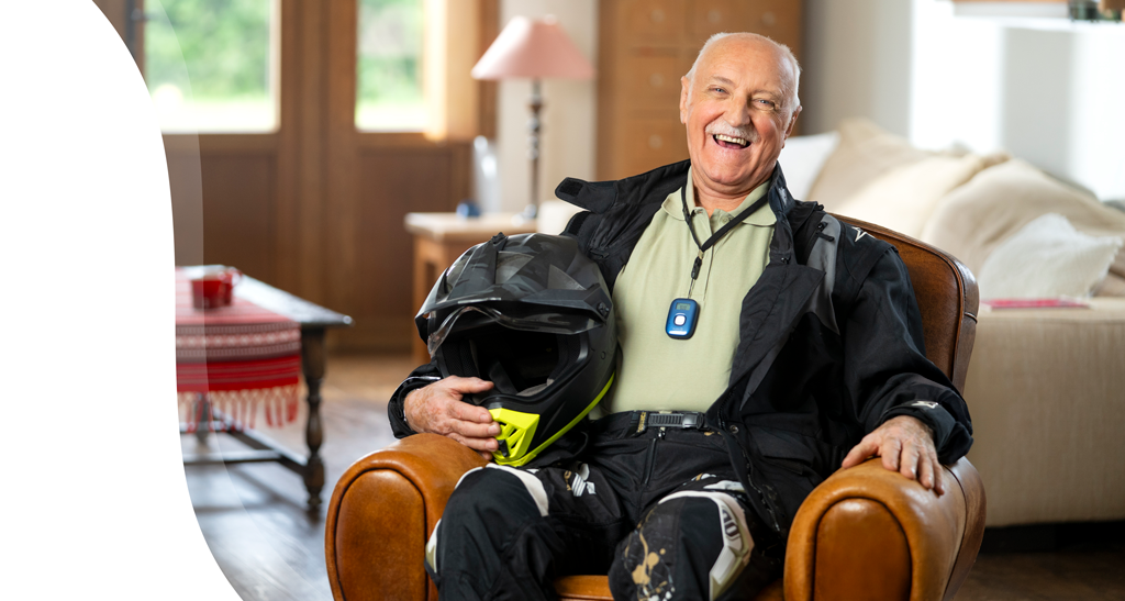 Oudere man zit lachend in een gezellige woonkamer, houdt een motorhelm vast en draagt een zwart motorpak met een medisch alarmapparaat om zijn nek.